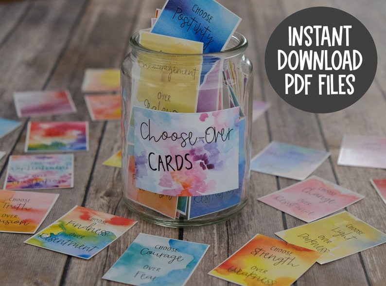 May include: A glass jar filled with colourful cards featuring watercolour designs. The cards have inspirational messages such as "Choose Positivity" and "Choose Courage over Fear". The jar is labelled "Choose-Over Cards".