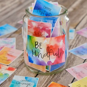 May include: A glass jar filled with colourful cards with watercolour backgrounds and inspirational messages. The jar has a label that says "BE Mindful".