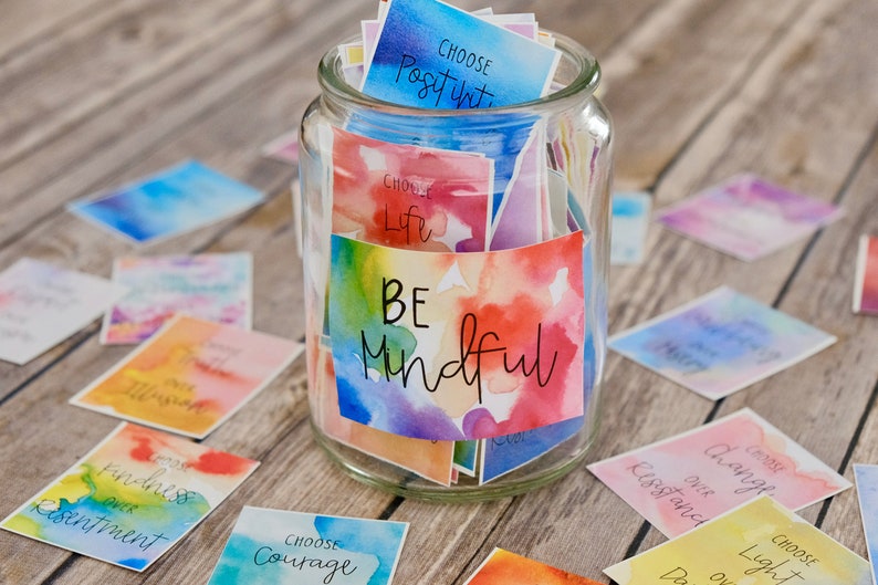 May include: A glass jar filled with colourful cards with watercolour backgrounds and inspirational messages. The jar has a label that says "BE Mindful".