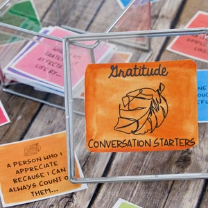 K&ouml;nnte beinhalten: Eine transparente Glasbox gef&uuml;llt mit kleinen orangefarbenen, rosa und blauen Karten mit handschriftlichem Text. Die Box ist mit "Gratitude Conversation Starters" beschriftet.