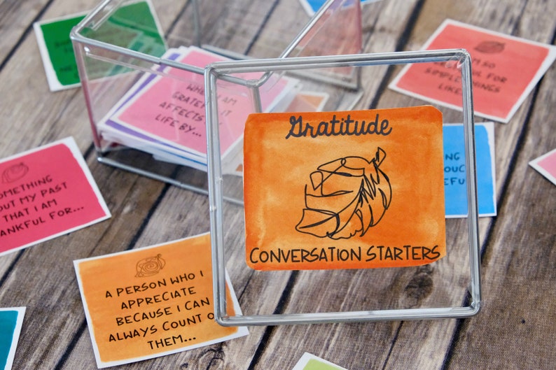 K&ouml;nnte beinhalten: Eine transparente Glasbox gef&uuml;llt mit kleinen orangefarbenen, rosa und blauen Karten mit handschriftlichem Text. Die Box ist mit "Gratitude Conversation Starters" beschriftet.