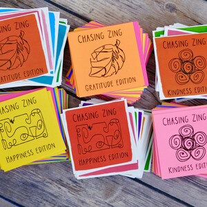 K&ouml;nnte beinhalten: Sechs Stapel bunter Karten mit dem Text "Chasing Zing" und verschiedenen Editionsnamen: Gratitude, Happiness und Kindness. Jeder Stapel hat eine andere Farbe und ein einzigartiges Grafikdesign.