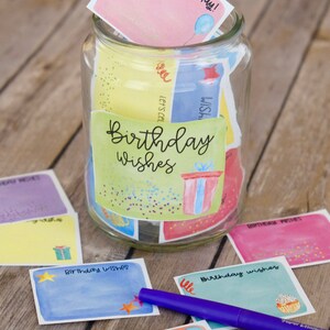 May include: A glass jar filled with colorful cards that say "Birthday Wishes" on them. The cards have different designs, including a gift box, a balloon, and a cupcake. There is a purple pen next to the jar.