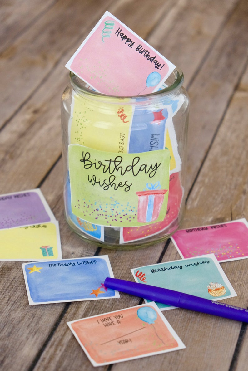 May include: A glass jar filled with colorful cards that say "Birthday Wishes" on them. The cards have different designs, including a gift box, a balloon, and a cupcake. There is a purple pen next to the jar.