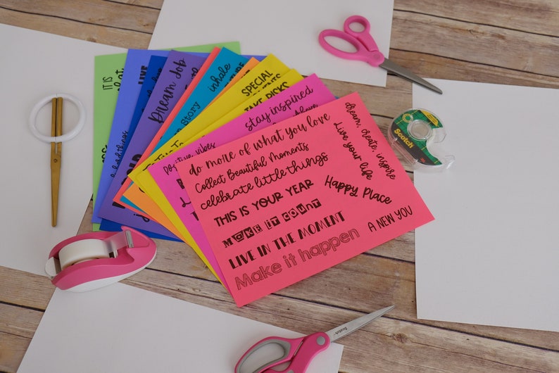 Peut inclure: Une pile de cartes en papier de couleur avec des citations inspirantes &eacute;crites &agrave; l'encre noire. Les cartes sont dispos&eacute;es en &eacute;ventail sur une surface en bois. On voit &eacute;galement des ciseaux, du ruban adh&eacute;sif et d'autres fournitures d'artisanat sur l'image.