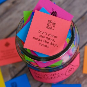 May include: A glass jar filled with colorful paper squares, each with an inspirational message. The top square is red and reads "Don't count the days, make the days count."