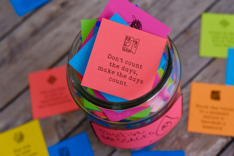 May include: A glass jar filled with colorful paper squares, each with an inspirational message. The top square is red and reads "Don't count the days, make the days count."