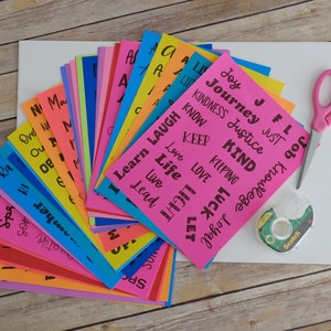 Peut inclure: Un ensemble de cartes en papier color&eacute;es avec des mots positifs imprim&eacute;s dessus, tels que "Joie", "Bienveillant", "Apprendre", "Amour" et "Vie". Les cartes sont dispos&eacute;es en &eacute;ventail sur une surface blanche. On voit &eacute;galement des ciseaux, du ruban adh&eacute;sif et une feuille de papier blanche sur l'image.