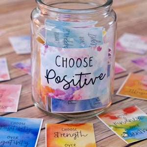 May include: A glass jar filled with colourful square cards with watercolour backgrounds and black text. The cards have positive affirmations such as "Choose Positive", "Choose Strength over Weakness", and "Choose Kindness over Resentment".