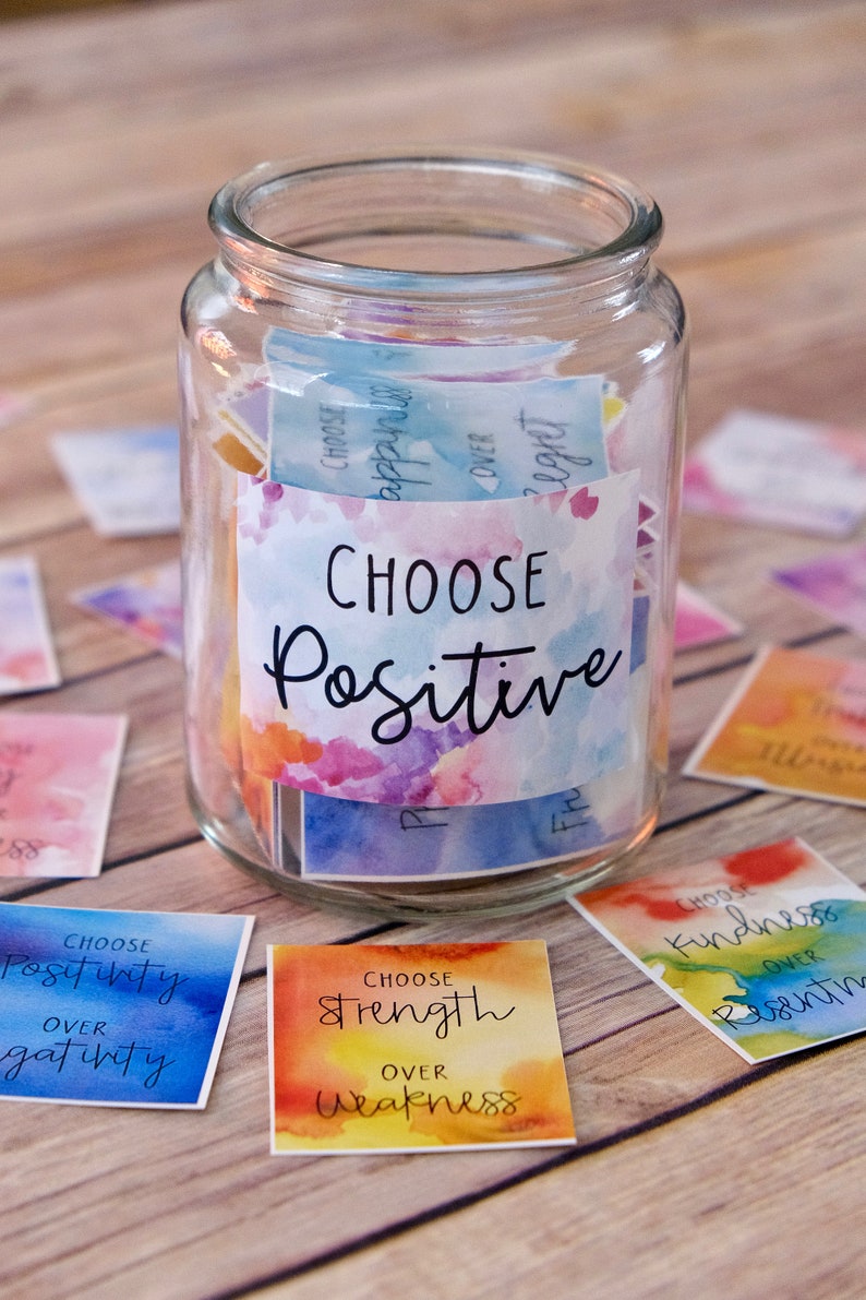 May include: A glass jar filled with colourful square cards with watercolour backgrounds and black text. The cards have positive affirmations such as "Choose Positive", "Choose Strength over Weakness", and "Choose Kindness over Resentment".