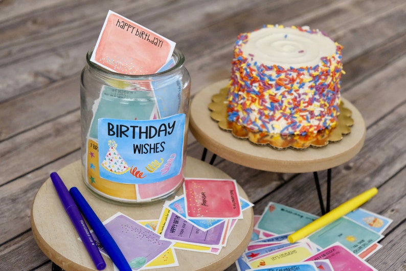 May include: A glass jar filled with colorful cards that say "Birthday Wishes" on the label. The jar is on a wooden surface with a cake and other cards. The cards have different birthday wishes written on them.