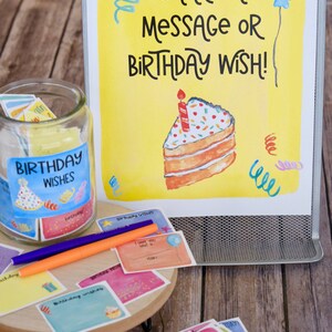 May include: A yellow sign with the text "SHARE A MESSAGE OR BIRTHDAY WISH!" and a drawing of a slice of cake with a candle. A glass jar with a label that says "BIRTHDAY WISHES" is filled with colorful paper slips. There are also colorful paper slips scattered on a wooden surface.
