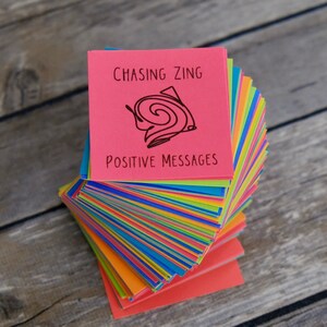 May include: A stack of colorful sticky notes with a pink note on top that says "Chasing Zing Positive Messages" with a doodle of a swirl on it.