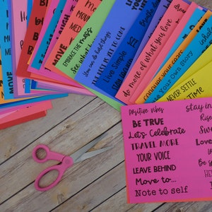 Peut inclure: Une pile de cartes color&eacute;es avec des citations et des phrases inspirantes, telles que "Soyez vrai", "C&eacute;l&eacute;brons", "Voyagez plus", "Votre voix", "Laissez derri&egrave;re", "D&eacute;placez-vous vers...", "Note &agrave; soi", "Restez inspir&eacute;", "S'&eacute;lever au-dessus", "Belle vie", "Amour et famille", "Cherchez...", "Soyez spontan&eacute;", "Aidez les autres", "Je t'aime", "Vibrations positives", "Embrassez le d&eacute;sordre", "Voyez ce que vous pouvez faire", "Vivez simplement", "Le meilleur est &agrave; venir", "Faites plus de ce que vous aimez", "Conqu&eacute;rir vos peurs", "&Eacute;crivez votre propre histoire", "Ne vous installez jamais", "Temps", "Inspirez", "Sp&eacute;cial", "Prenez des risques", "Choisissez la joie", "C&oelig;ur et &acirc;me", "Soyez pr&eacute;sent", "Amour et", "Pause", "Bougez", "Respirez profond&eacute;ment", "R&ecirc;ve", "Rire", "M&eacute;d", "&Eacute;quipe", "Ra", "A", "Th835", "110", "N", "I", "R", "M", "B", "( ", "Nous", "H". Une paire de ciseaux roses est au premier plan.