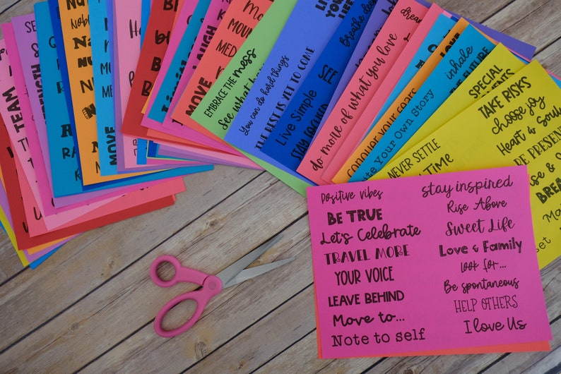 Peut inclure: Une pile de cartes color&eacute;es avec des citations et des phrases inspirantes, telles que "Soyez vrai", "C&eacute;l&eacute;brons", "Voyagez plus", "Votre voix", "Laissez derri&egrave;re", "D&eacute;placez-vous vers...", "Note &agrave; soi", "Restez inspir&eacute;", "S'&eacute;lever au-dessus", "Belle vie", "Amour et famille", "Cherchez...", "Soyez spontan&eacute;", "Aidez les autres", "Je t'aime", "Vibrations positives", "Embrassez le d&eacute;sordre", "Voyez ce que vous pouvez faire", "Vivez simplement", "Le meilleur est &agrave; venir", "Faites plus de ce que vous aimez", "Conqu&eacute;rir vos peurs", "&Eacute;crivez votre propre histoire", "Ne vous installez jamais", "Temps", "Inspirez", "Sp&eacute;cial", "Prenez des risques", "Choisissez la joie", "C&oelig;ur et &acirc;me", "Soyez pr&eacute;sent", "Amour et", "Pause", "Bougez", "Respirez profond&eacute;ment", "R&ecirc;ve", "Rire", "M&eacute;d", "&Eacute;quipe", "Ra", "A", "Th835", "110", "N", "I", "R", "M", "B", "( ", "Nous", "H". Une paire de ciseaux roses est au premier plan.