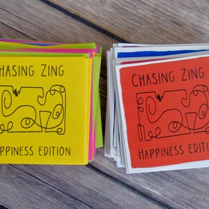 K&ouml;nnte beinhalten: Zwei Stapel Karten mit dem Text "Chasing Zing Happiness Edition" auf der Vorderseite. Der obere Kartenstapel ist gelb und der untere Stapel ist rot.