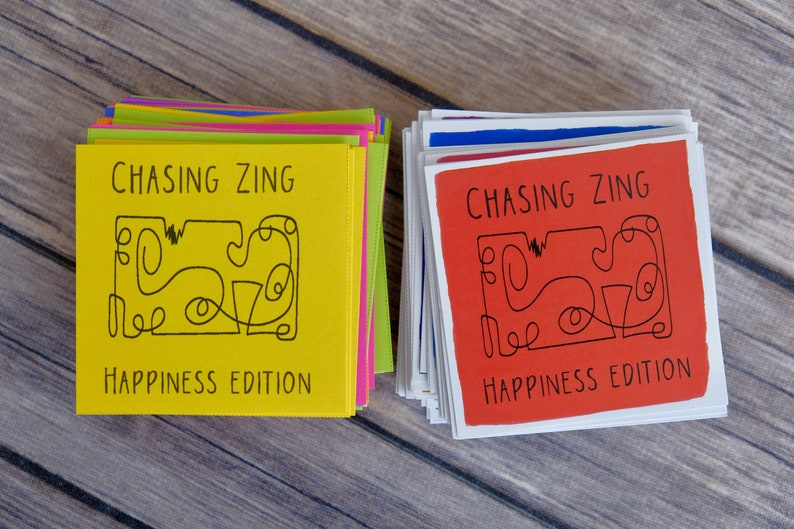 K&ouml;nnte beinhalten: Zwei Stapel Karten mit dem Text "Chasing Zing Happiness Edition" auf der Vorderseite. Der obere Kartenstapel ist gelb und der untere Stapel ist rot.