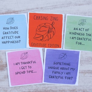 K&ouml;nnte beinhalten: F&uuml;nf bunte Karten mit einem Blattdesign und Text &uuml;ber Dankbarkeit. Die Karten lauten: "Wie wirkt sich Dankbarkeit auf unser Gl&uuml;ck aus?", "Chasing Zing Gratitude Edition", "Eine Handlung der Freundlichkeit, f&uuml;r die ich dankbar bin...", "Ich bin dankbar, dass ich Zeit verbringen kann...", und "Etwas Einzigartiges an meiner Familie, f&uuml;r das ich dankbar bin?"