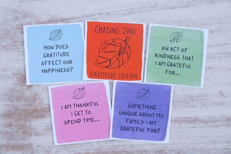 K&ouml;nnte beinhalten: F&uuml;nf bunte Karten mit einem Blattdesign und Text &uuml;ber Dankbarkeit. Die Karten lauten: "Wie wirkt sich Dankbarkeit auf unser Gl&uuml;ck aus?", "Chasing Zing Gratitude Edition", "Eine Handlung der Freundlichkeit, f&uuml;r die ich dankbar bin...", "Ich bin dankbar, dass ich Zeit verbringen kann...", und "Etwas Einzigartiges an meiner Familie, f&uuml;r das ich dankbar bin?"