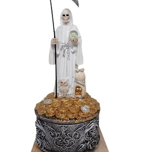 Peut inclure: Figurine blanche représentant une figure squelettique en robe à capuche, tenant une faux et une boule de cristal. La figurine est posée sur un coffre au trésor débordant de pièces d'or. Un hibou et un sac de pièces sont également présents.