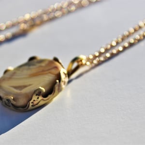 May include: A gold chain necklace with a pendant featuring a light brown agate stone set in a gold filigree frame.