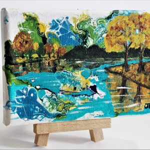 May include: Abstract painting on canvas with a blue, green, and yellow color scheme. The painting depicts a landscape with trees and water. The painting is on a small easel.