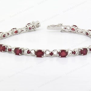 May include: A silver bracelet with a row of oval-shaped red gemstones. The bracelet has a delicate, intricate design.