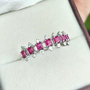 May include: A silver ring with a row of rectangular, bright red gemstones, set between smaller, clear stones. The ring is displayed in a maroon jewellery box with a white interior, highlighting its elegant design. The ring's design is a continuous band of alternating stones.