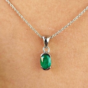 Puede incluir: Un collar de plata con un pequeño colgante de esmeralda de forma ovalada. La piedra preciosa verde brillante está engastada en un bisel de plata y cuelga de una delicada cadena de plata. El collar se muestra sobre un fondo de tono de piel.