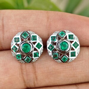 Pode incluir: Um par de brincos redondos com uma pedra preciosa central verde esmeralda, rodeada por pedras verdes e claras menores. Os brincos têm uma moldura de metal prateado e um design floral.