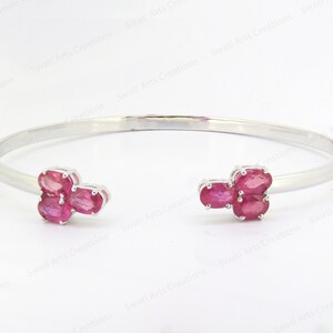 Può includere: Un bracciale a polsino aperto color argento con grappoli di pietre preziose rosa vivaci taglio ovale a ciascuna estremità. Le pietre preziose sono incastonate in metallo argentato. Il bracciale è esposto su sfondo bianco.