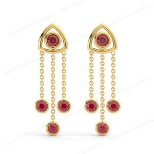 May include: Gold earrings with a triangular design and red gemstones. The earrings have three dangling chains with red gemstones at the end of each chain.