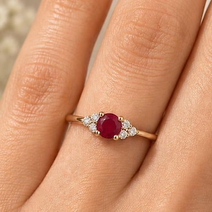 May include: A gold ring featuring a central oval ruby gemstone, flanked by smaller, clear gemstones. The ring is set on a delicate gold band, and is displayed on a person's hand. The background is softly blurred, with white flowers.