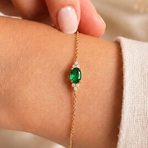 May include: A delicate gold bracelet featuring an oval emerald gemstone, flanked by two smaller white gemstones. The bracelet is on a slender gold chain, and is worn on a wrist.