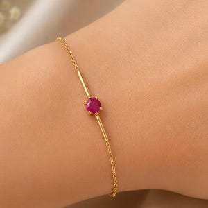 Può includere: Un delicato bracciale a catena in oro con una singola pietra preziosa rotonda di un vivido colore rosso rosato, montata su una montatura a griffe in oro e affiancata da due piccole barrette dorate su entrambi i lati della pietra.