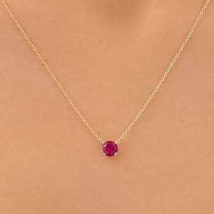 May include: A delicate gold chain necklace featuring a single, small, round-cut vibrant pinkish-red gemstone pendant centred on the chain. The necklace is worn around the neck, showcasing the minimalist design of the metal link chain.