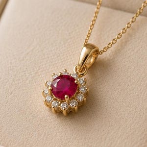 Könnte beinhalten: Eine Goldhalskette mit einem runden, tiefroten Rubinanhänger, der von einem Kranz aus kleinen, klaren, rund geschliffenen Diamanten umgeben ist. Der Anhänger hängt an einer feinen Goldkette und liegt in einer beigen, mit Samt ausgekleideten Schmuckschatulle.