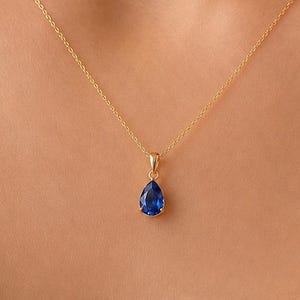 Könnte beinhalten: Eine zarte Goldkettenhalskette mit einem einzelnen birnenförmigen blauen Saphiranhänger. Der Anhänger ist in einer Goldfassung befestigt und hängt an einer dünnen, polierten Goldkette, die am Schlüsselbeinbereich aufliegt.