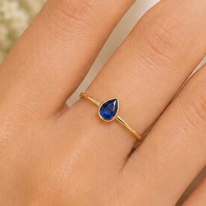 Op de afbeelding: Een dunne gouden ring met een enkele peervormige blauwe saffier in een gladde zetting. De ring heeft een gladde, gepolijste gouden band die om de vinger zit en de diepblauwe kleur van de druppelvormige edelsteen benadrukt.