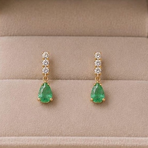 Puede incluir: Un par de pendientes colgantes de oro con dos pequeñas piedras redondas transparentes colocadas verticalmente sobre una piedra verde en forma de pera. Los pendientes se presentan dentro de un joyero de terciopelo beige con tapa.