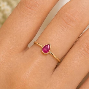 Op de afbeelding: Een verfijnde gouden ring met een enkele peervormige, felroze edelsteen in een gladde zetting. De dunne, gepolijste gouden band omsluit de vinger en laat de druppelvormig geslepen steen zien als het centrale middelpunt van het sieraad.