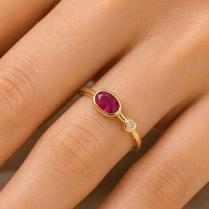 May include: A gold ring featuring an oval-shaped, deep pinkish-red gemstone set in a bezel, accompanied by a smaller, round, clear white gemstone set next to it on a thin, polished gold band that encircles the finger.
