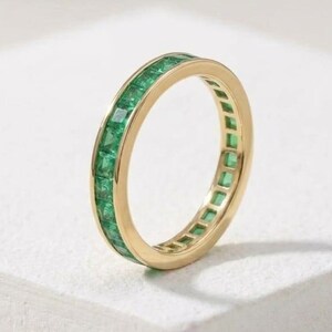 Op de afbeelding: Een gouden ring met een band van vierkant geslepen smaragden. De ring staat op een wit oppervlak, waardoor de levendige groene edelstenen en de gepolijste gouden band worden benadrukt. De ring is een sieraad.