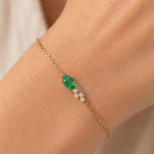 May include: A delicate gold chain bracelet featuring a central oval-cut green gemstone flanked by two small, round, clear white stones. The bracelet is secured with a fine link chain, creating a subtle and refined piece of jewelry for the wrist.
