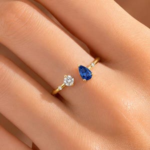 Könnte beinhalten: Ein offener Goldring mit einem birnenförmigen blauen Saphir an einem Ende und einem runden weißen Diamanten im Brillantschliff am anderen Ende, akzentuiert durch zwei winzige weiße Diamanten, die beidseitig in die dünne Goldschiene gefasst sind.