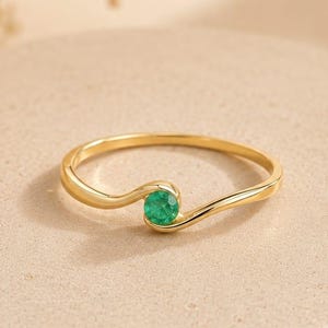 Puede incluir: Un delicado anillo de oro con un diseño ondulado, con una pequeña gema redonda de color verde esmeralda. El anillo se exhibe sobre una superficie texturizada de tono neutro, mostrando su elegante simplicidad. La banda del anillo es delgada y pulida, resaltando el verde vibrante de la gema.