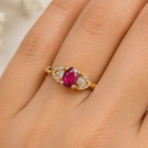 May include: A gold ring featuring a central oval-cut vibrant pinkish-red gemstone flanked by two small clear accent stones on each side, set within a delicate gold band with leaf-like metal detailing surrounding the stones.
