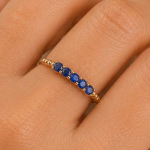 Peut inclure: Une bague en or ornée d'une rangée de cinq petites pierres précieuses rondes d'un bleu éclatant, serties horizontalement le long de l'anneau. La bande métallique présente un design perlé délicat et texturé qui encadre les pierres.