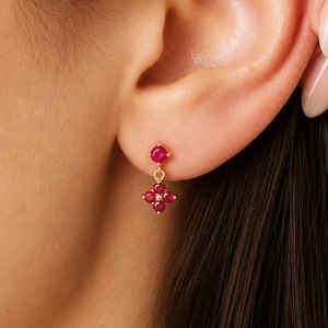 Peut inclure: Une boucle d'oreille pendante en or composée d'une petite pierre rouge ronde en haut, reliée par une fine chaîne en or à un motif floral à quatre pétales formé de pierres rouges rondes. La boucle d'oreille est portée au lobe.