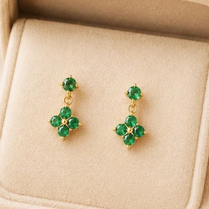 May include: A pair of gold drop earrings featuring a small round green gemstone at the top and a four-leaf clover shape composed of four round green gemstones at the bottom, displayed inside a beige velvet jewelry box with a soft fabric lining.